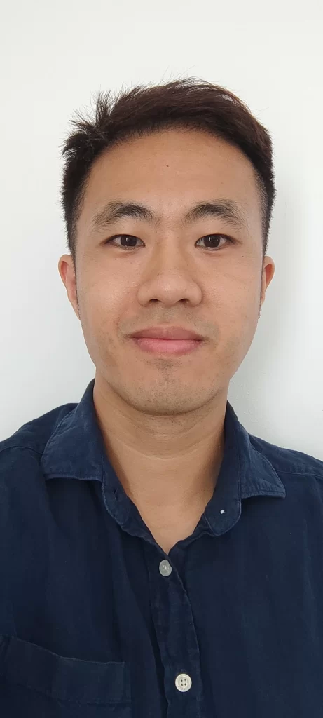 a closeup headshot of an asian man with short black hair and dark brown eyes, he is wearing a dark blue button-up collared shirt and is smiling slightly at the camera against a plain white background