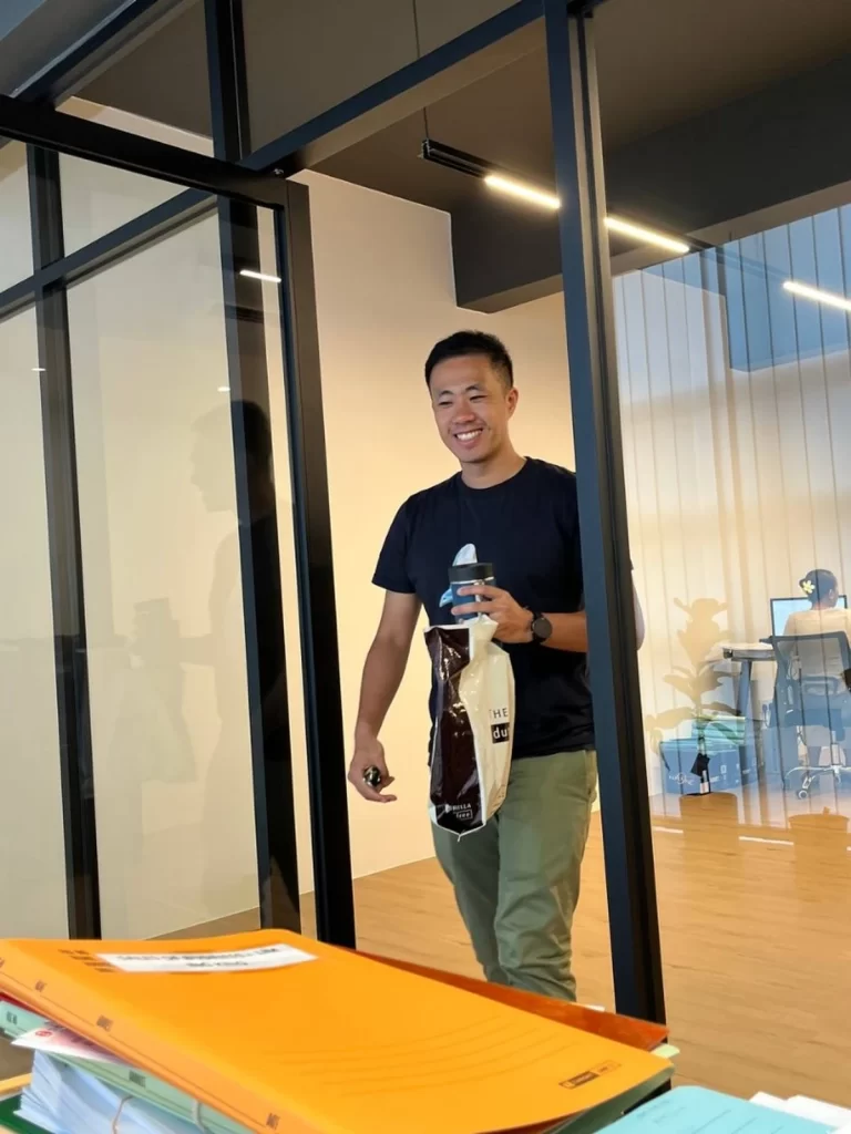 a smiling asian man in a black t-shirt and green pants walks through a modern glass and black-framed office door, he is carrying a water bottle and a bag of coffee beans from dutch colony coffee co
