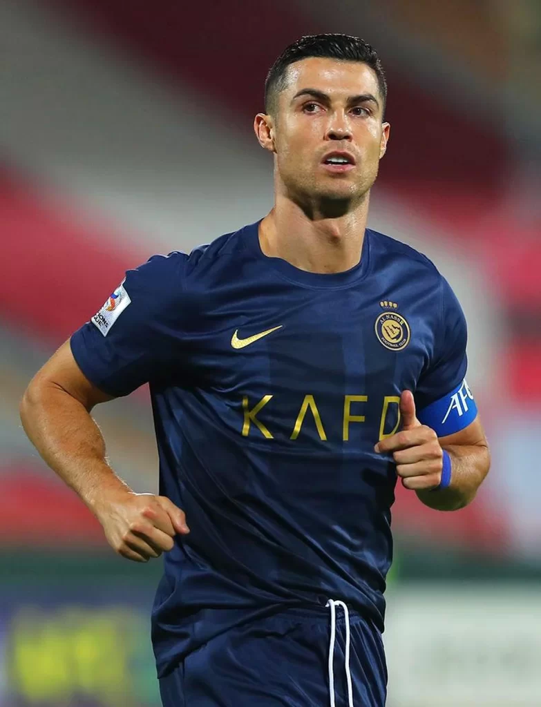 soccer player cristiano ronaldo jogs on the field, wearing a navy blue al-nassr fc jersey with gold lettering from nike and a blue captain's armband, he has a focused expression