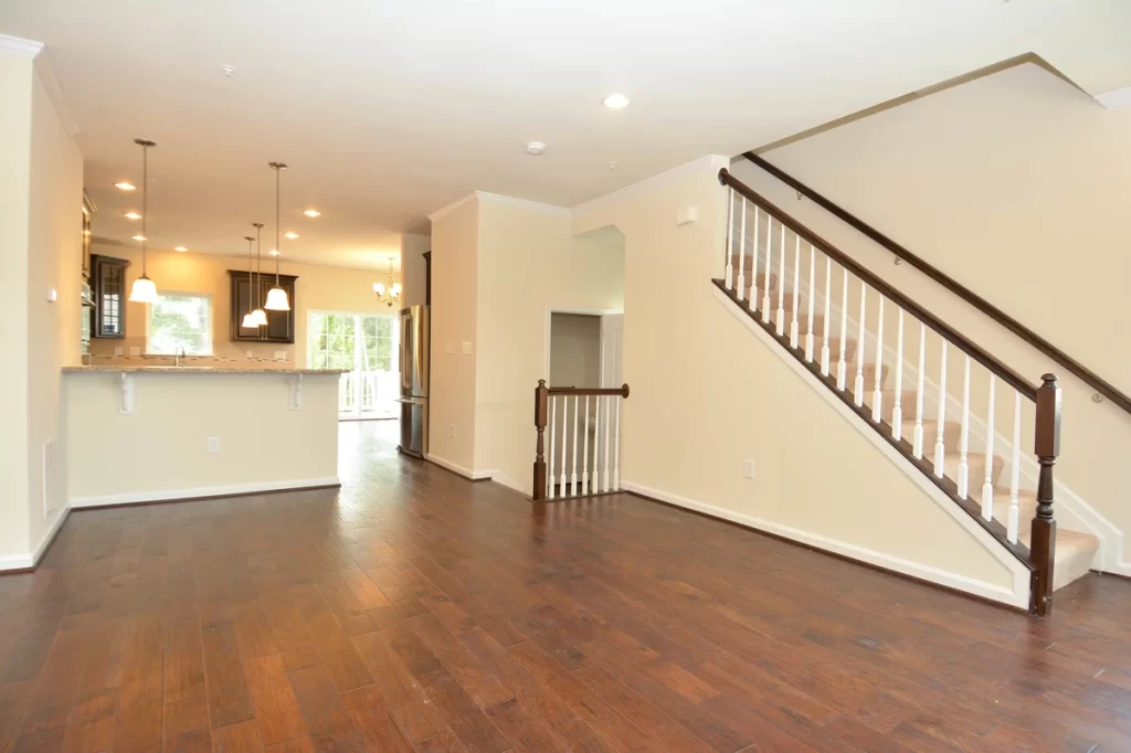 a spacious, empty room with dark brown hardwood floors, beige walls, and a staircase with a dark wood railing and white spindles, in the background, a kitchen with dark cabinets and a breakfast bar is visible