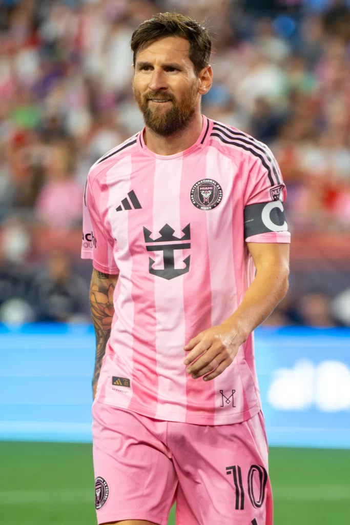 soccer player lionel messi walks on the field with a slight smile, he has a beard and is wearing the pink and black striped inter miami cf jersey with the adidas logo and a captain's armband