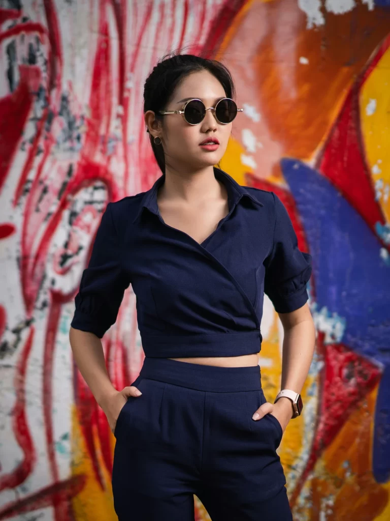 an asian woman with her hair tied back wears round sunglasses and a navy blue two-piece outfit, she stands with her hands in her pockets in front of a colorful, abstract graffiti wall