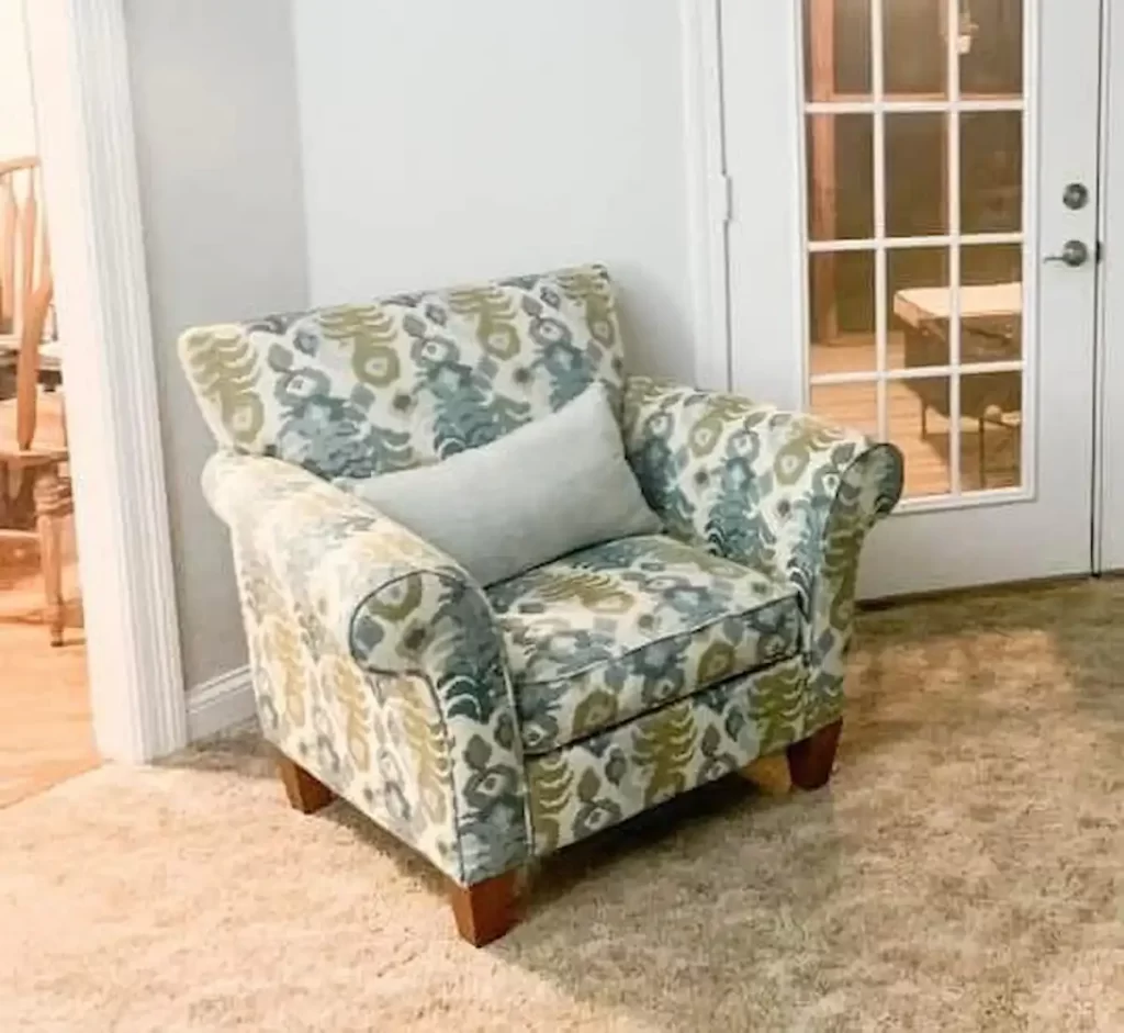 a comfortable-looking armchair with a blue, green, and cream floral damask pattern, it has rolled arms, dark wood legs, and a light grey lumbar pillow, sitting on a beige carpet next to a white door