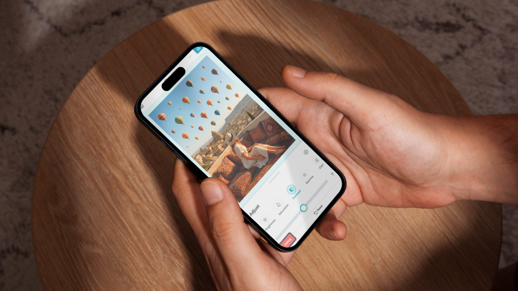 overhead shot of hands holding a smartphone over a light wooden table, the phone screen displays the image adjustment interface with a photo of a woman sitting on a terrace with hot air balloons in the background, the contrast setting is selected on the bottom slider menu which also includes options for brightness saturation and gamma