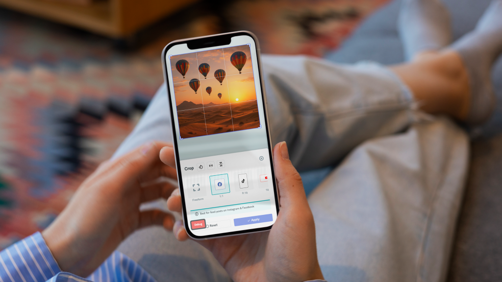 close up view of a person holding a smartphone displaying the crop tool within the image editor interface, the screen shows a photo of hot air balloons over a desert landscape with grid lines overlaying the image, crop options for freeform and square aspect ratios are visible at the bottom along with a tip for instagram and facebook posts, the person wears a blue striped shirt and jeans against a blurred living room background