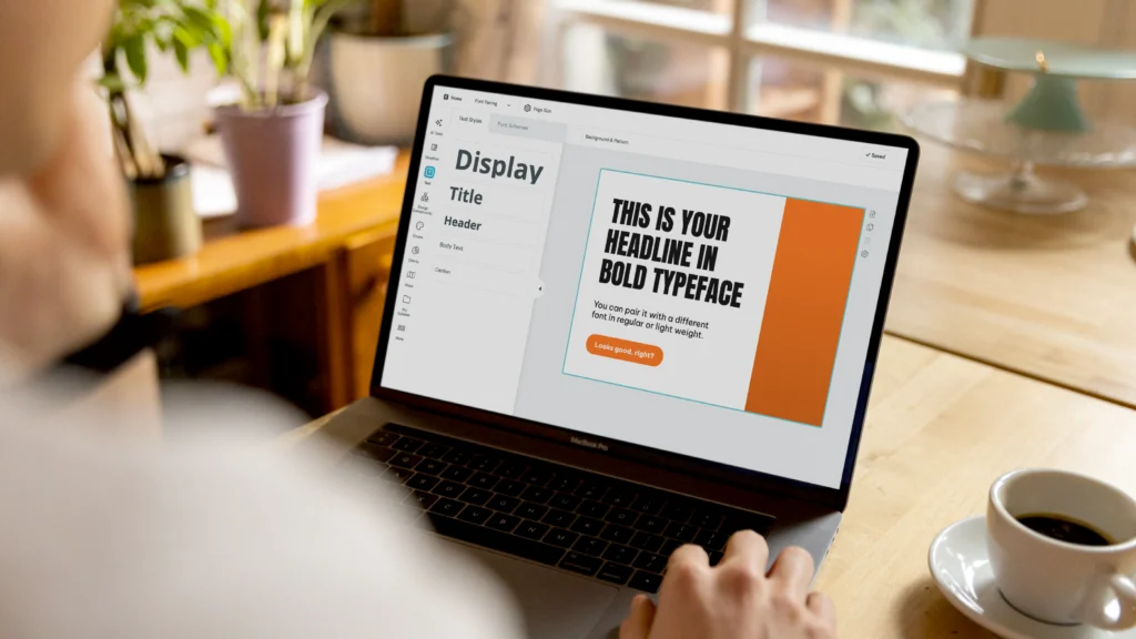 a person sits at a wooden desk with a cup of coffee, working on a laptop that displays a design interface with text editing options, the screen shows a bold headline and a button, the overall scene is warm and professional