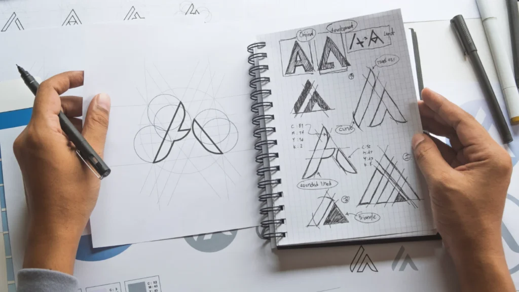 two hands hold and draw in an open spiral notebook filled with ink sketches of the letter a in triangular and mountain-inspired forms, with color values and design notes written alongside, while the left hand sketches geometric circle-based shapes on a separate loose sheet surrounded by additional logo printouts