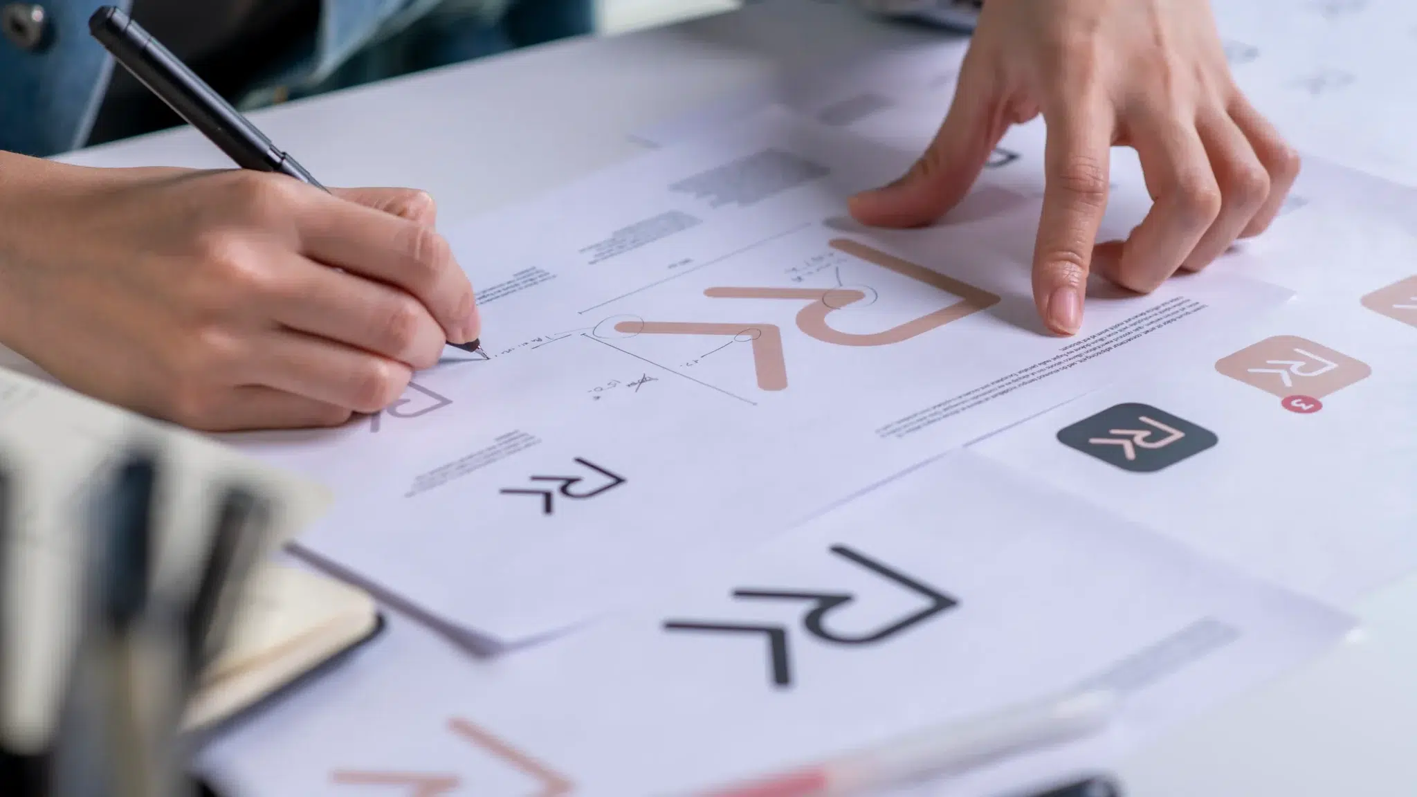 two hands examine a collection of printed brand identity sheets spread across a white desk, featuring a stylized two-letter monogram in both a thin black line version and a rounded rose gold version, with one sheet showing the logo inside a dark rounded app icon and another displaying dimension guidelines and spacing annotations