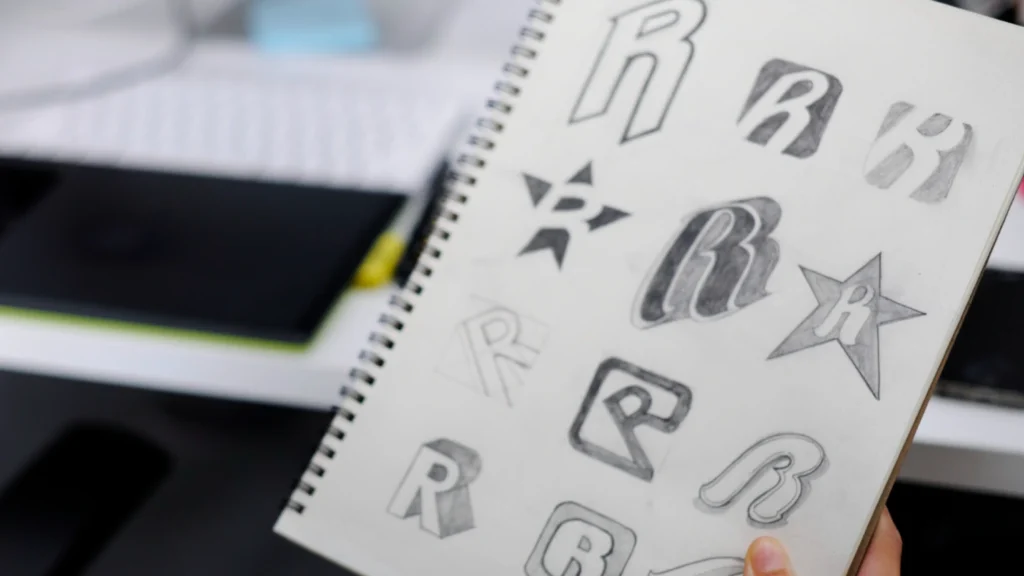 a hand holds an open spiral sketchbook displaying multiple pencil sketches of the letter r in various styles, including bold block letters, three-dimensional renderings, a star-shaped emblem with an r, rounded bubbly letterforms, and shadowed serif designs, set against a blurred desk workspace in the background