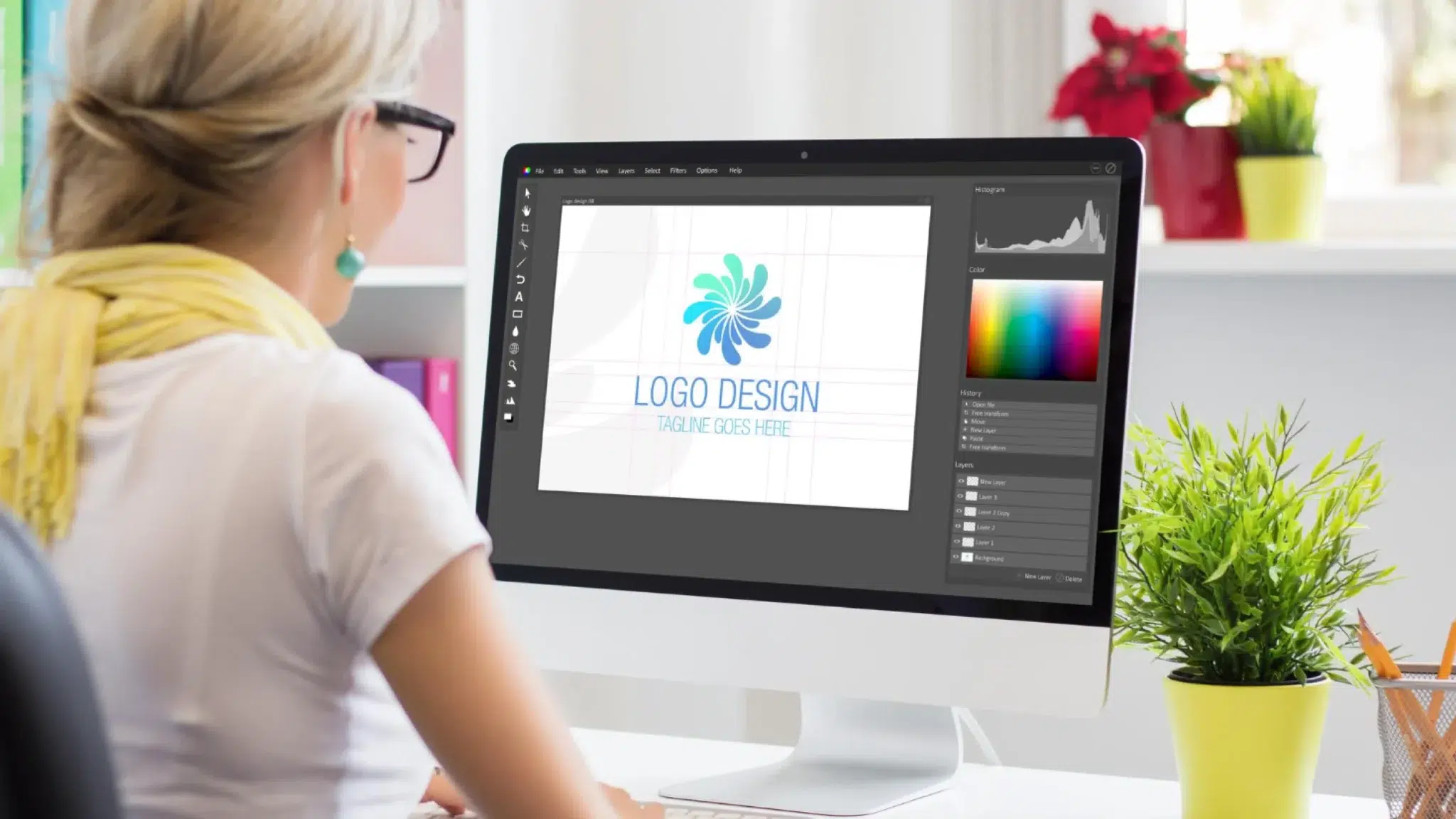 a woman with blonde hair pulled back and glasses resting on her head sits facing a large desktop monitor displaying a teal pinwheel-style graphic with the placeholder text logo design and tagline goes here in a design application with layers and color panels visible, surrounded by plants and colorful books on shelves, illustrating negative space