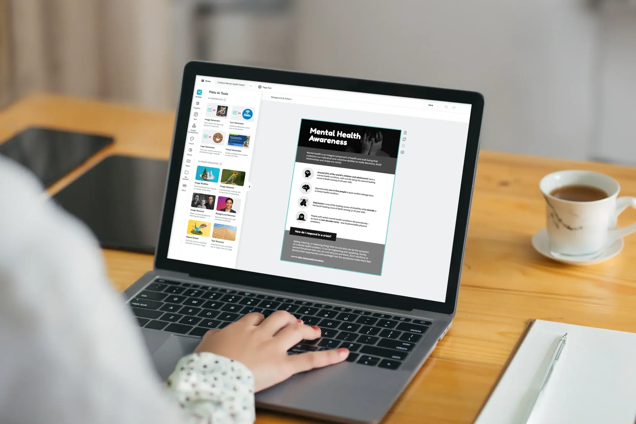 over the shoulder view of a person typing on a macbook displaying the pikto ai editor with a mental health awareness poster open in the workspace and the ai tools panel showing image generator, icon generator, logo generator, and visual generator alongside a white coffee cup and a notebook on a wooden desk
